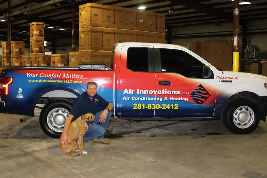 An Air Innovations owner kneeling beside a wrapped service pickup truck in the Cypress warehouse, with a dog